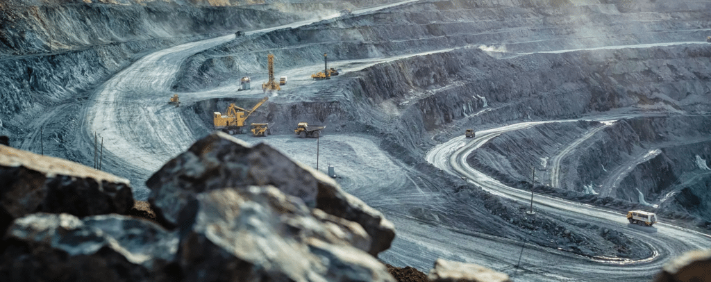 Open-pit mine with winding dirt roads, large excavators, and trucks working on stepped rocky slopes, surrounded by dust and loose rocks.