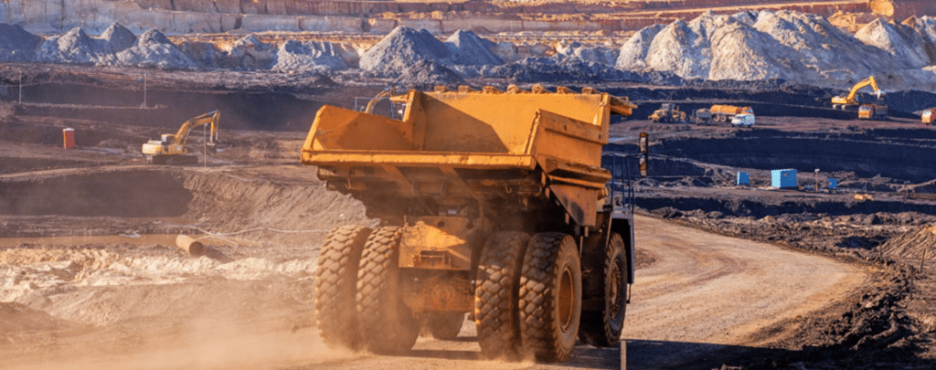 A large dump truck drives on a dirt road in an open-pit mining site, with excavators and piles of earth in the background.