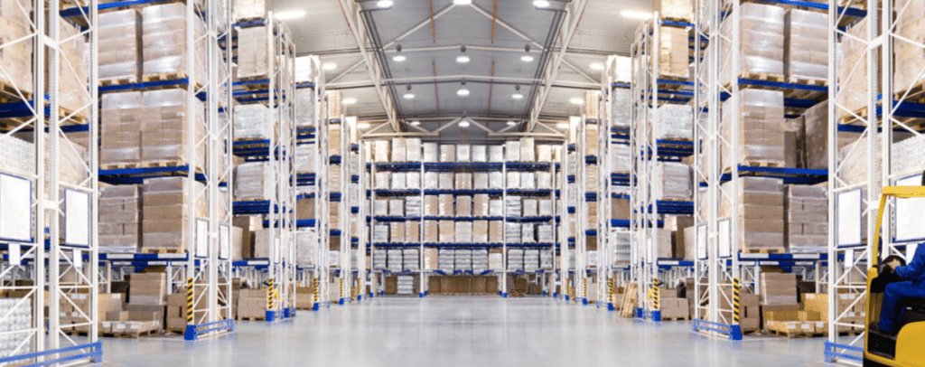 Large warehouse interior with tall shelves stacked with boxes and pallets, empty floor space in the foreground, and overhead lighting.