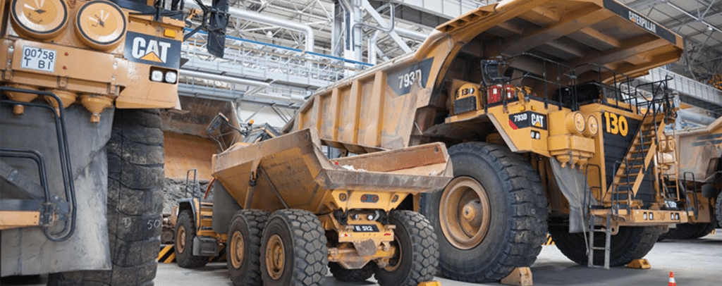 Large Caterpillar dump trucks and machinery are parked inside a spacious industrial facility with high ceilings and exposed pipes.