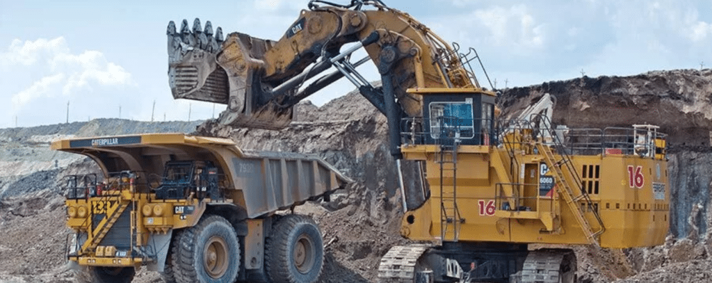 A large excavator loads soil into a dump truck at an open-pit mining site under a partly cloudy sky.