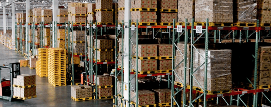 Large warehouse with tall racks holding stacked pallets of boxes, a forklift, and organized rows of goods ready for distribution.