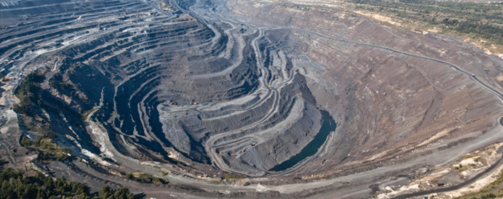 Aerial view of a large open-pit mine with spiraling terraced roads and exposed layers of earth and rock. Sparse vegetation surrounds the mine.