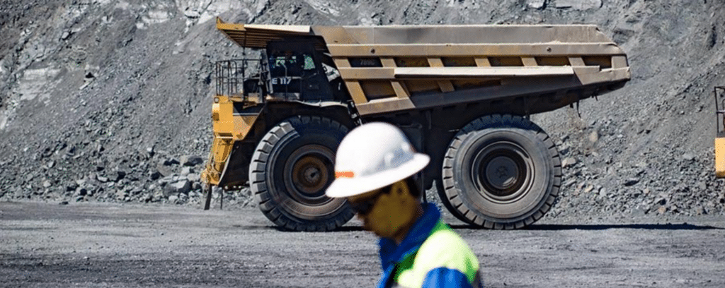 A large mining dump truck is parked on a gravel surface, with a worker in a hard hat and safety vest walking in the foreground.