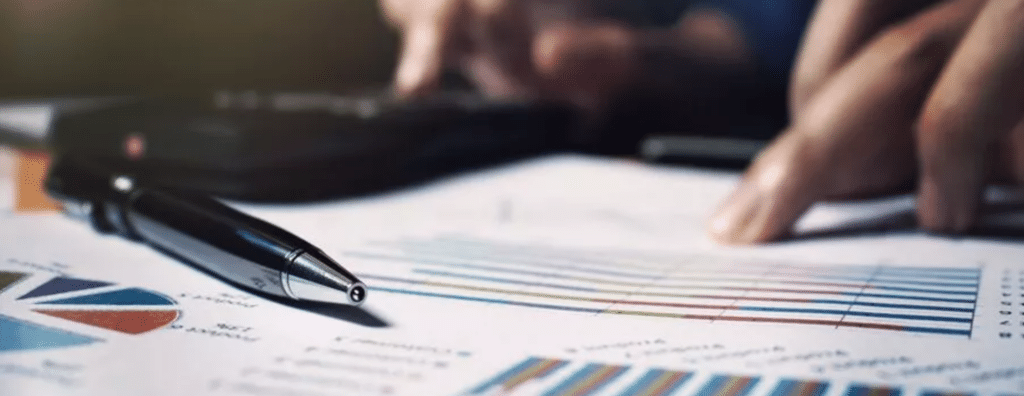 A pen rests on financial documents with charts and graphs, while a person uses a calculator in the background.