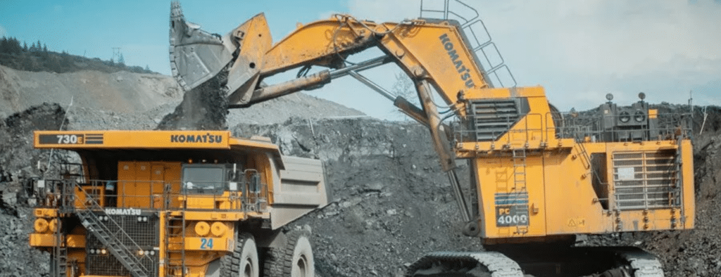 A large excavator loads soil into a Komatsu dump truck at a mining site, with rocky terrain and distant trees in the background.