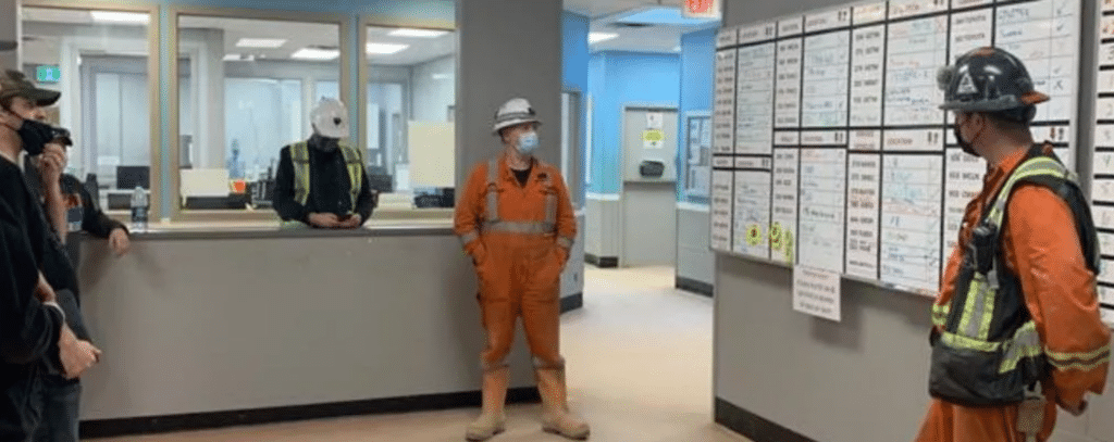 Several workers in safety gear and hard hats stand in an office, some facing a wall-mounted scheduling or tracking board.