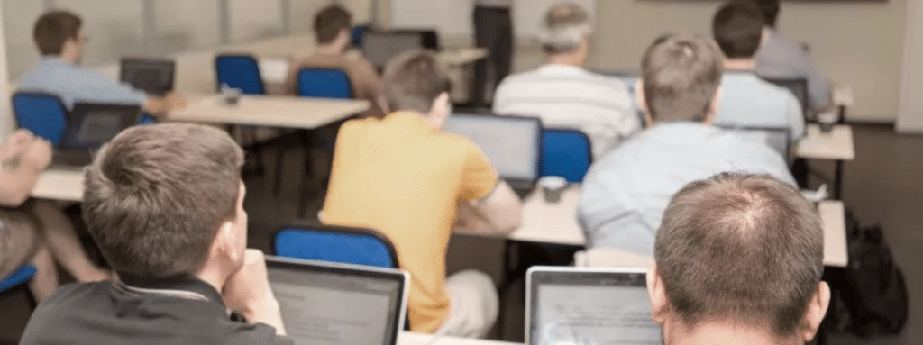 A group of people sit at desks with laptops, facing a presenter in a classroom or training room setting.