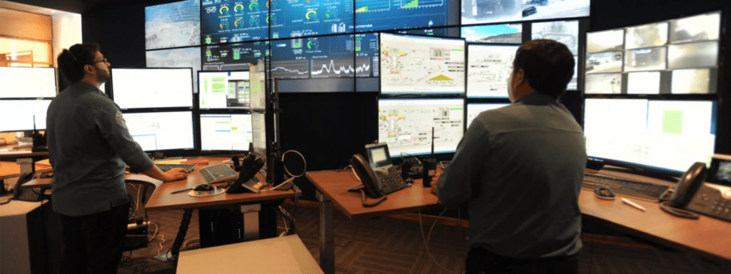 Two people stand in front of multiple computer monitors displaying data, graphs, and surveillance feeds in a control room.