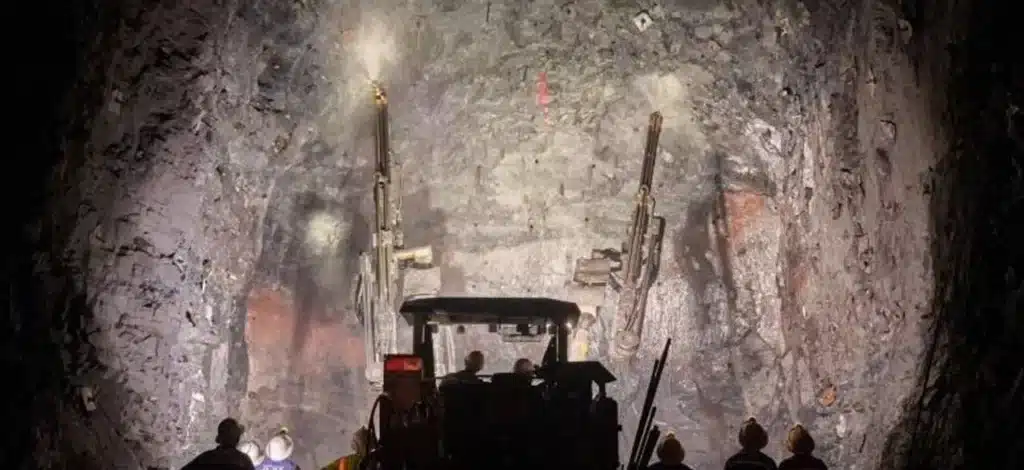 Workers operate machinery drilling into a rocky tunnel face, illuminated by bright lights inside an underground construction site.