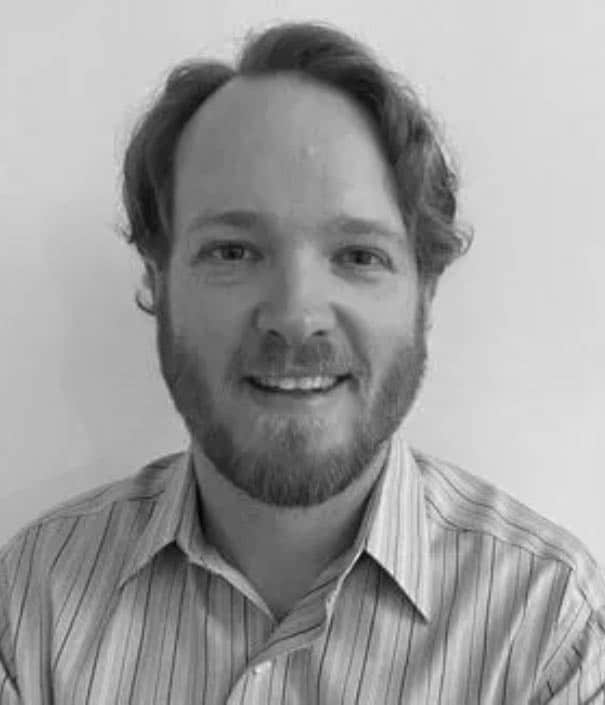 A man with a beard and wavy hair, wearing a striped button-up shirt, poses in front of a plain light background. The image is in black and white.