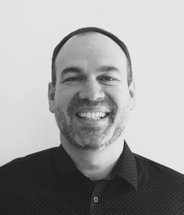 A man with short hair and a beard smiles at the camera, wearing a dark, collared shirt against a plain white background.