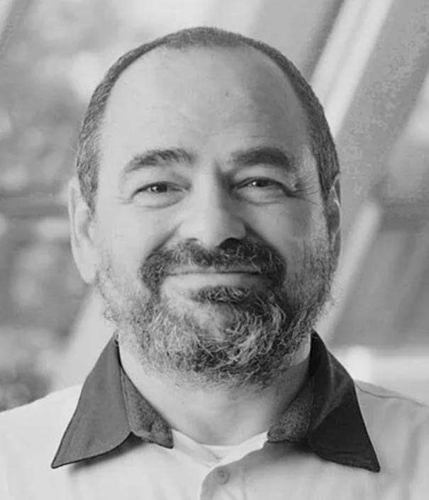A middle-aged man with a beard and mustache, smiling, wearing a collared shirt. The background is blurred and appears to be indoors with large windows.