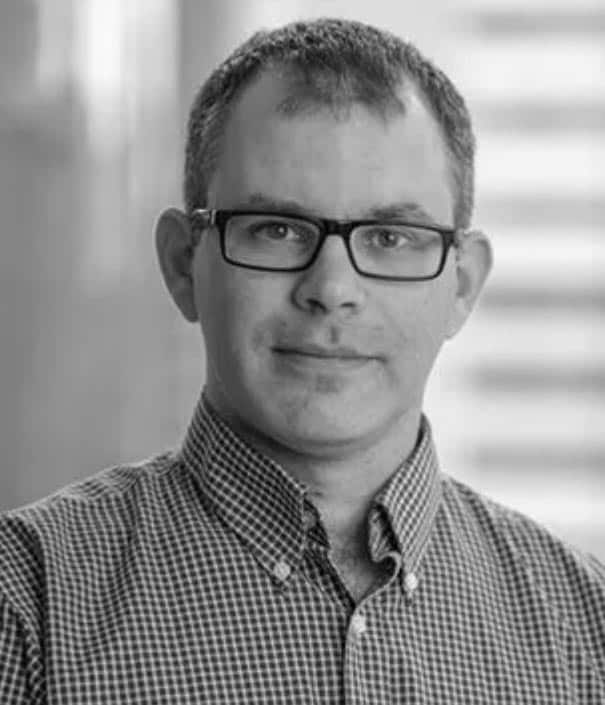 A man wearing glasses and a checkered button-up shirt looks at the camera, set against a blurred indoor background.
