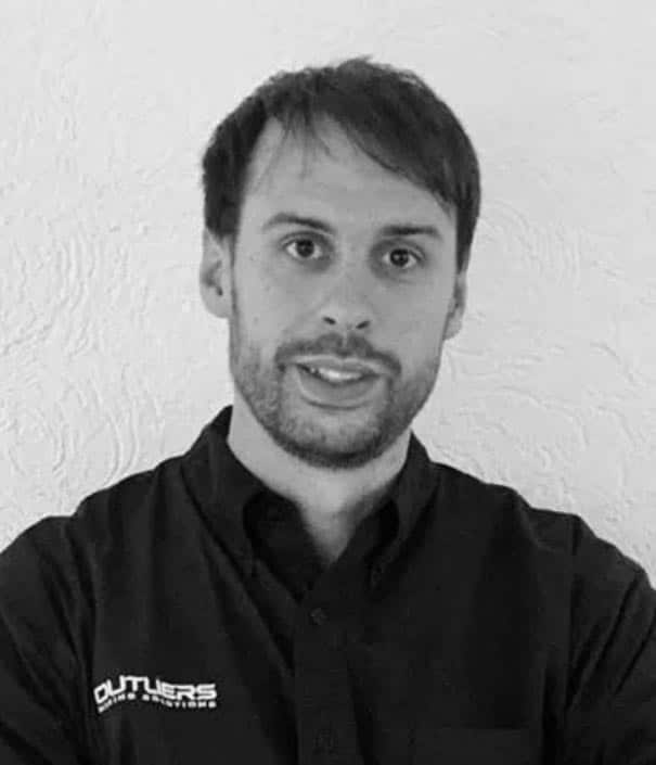 Man with short hair and beard wearing a dark shirt that reads “OUTLIERS” on the chest, standing in front of a textured white wall.