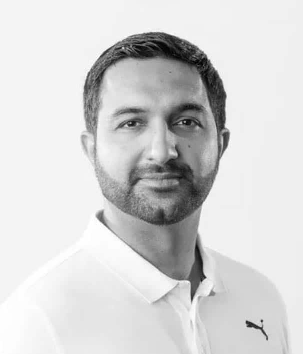 A man with short dark hair and a trimmed beard wears a light-colored collared shirt with a Puma logo, looking at the camera against a plain background.