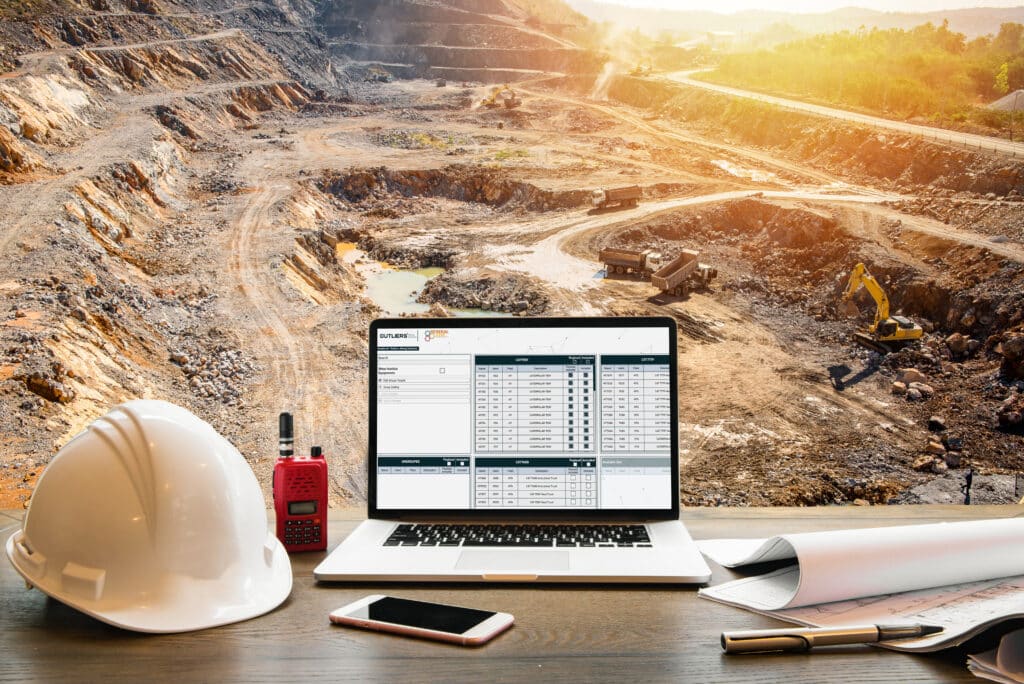 A construction site with excavators and trucks, viewed from a desk with a hard hat, laptop showing data, walkie-talkie, smartphone, and blueprints in the foreground.