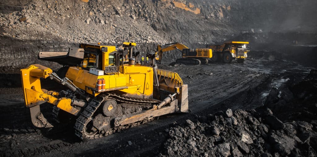 Large yellow bulldozer, excavator, and dump truck operate in an open-pit mining site, surrounded by dark soil and rock.