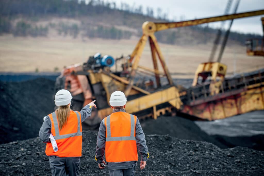 Two workers in orange safety vests and helmets observe and point towards large mining equipment at an open-pit mining site.