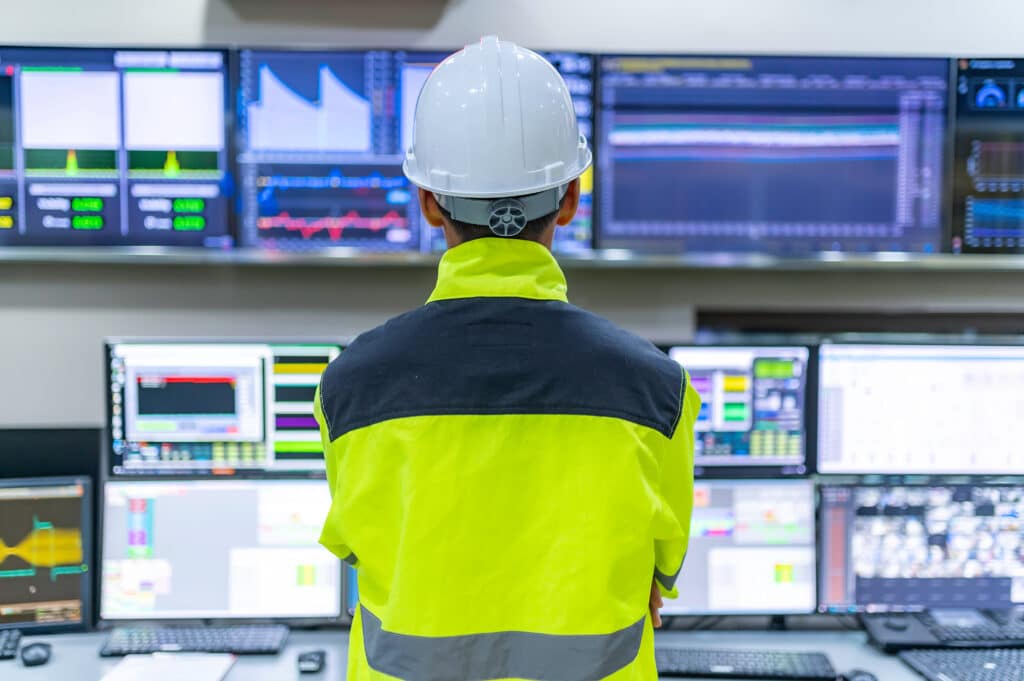 A person wearing a high-visibility jacket and hard hat monitors multiple screens displaying data and graphs in a control room.
