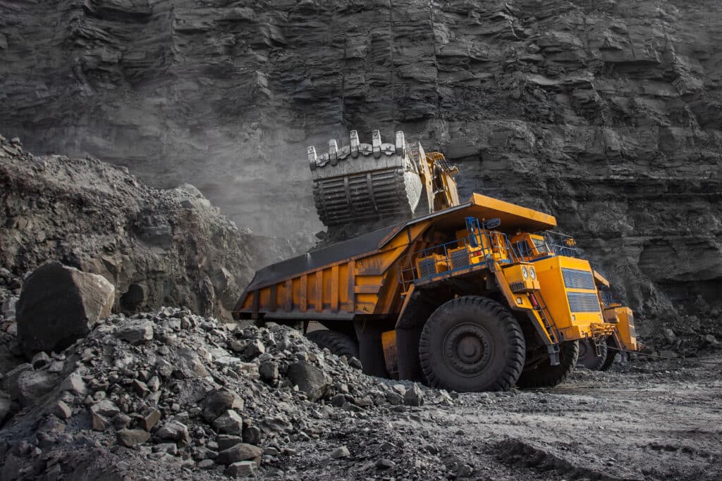 A large yellow mining truck is being loaded with rocks by an excavator in a rocky open-pit mine.