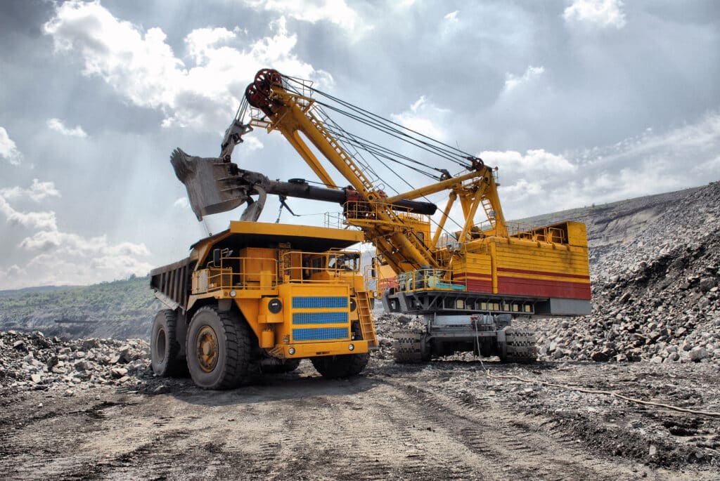 A large yellow excavator loads rocks into a dump truck at an open-pit mining site under a cloudy sky.