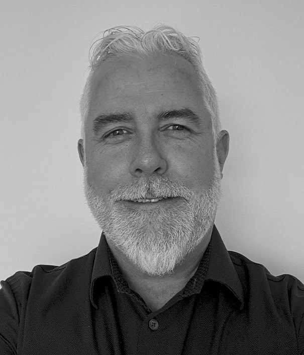 A middle-aged man with short white hair and a beard is smiling at the camera. He is wearing a dark collared shirt and is set against a plain background. The image is in black and white.